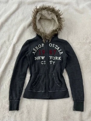 Mörkgrå Aeropostale hoodie med fuskpäls - Mörkgrå hoodie från Aeropostale med dragkedja, broderad text '1987 New York City' i vitt och rött på bröstet. Utrustad med huva fodrad med mjuk fuskpäls och fickor framtill. Perfekt för en chill och avslappnad stil. (lånade bilder)