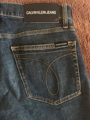 Blå jeans från Calvin Klein Jeans - Säljer ett par klassiska blå jeans från Calvin Klein Jeans med snygga kontrastsömmar och logga på bakfickan. Jeansen har fem fickor och normal midja. Materialet är slitstark denim i bomull. Perfekta för en avslappnad och stilren look.