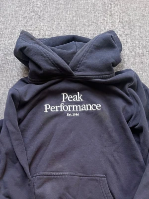 Svart hoodie Peak Performance - Den behövs tvättas så på bilden var den inte tvättat men tvättar såklart inan jag postar. 😊Snygg svart hoodie från Peak Performance med vit logga och text 'Est. 1986' på bröstet. Klassisk modell med stor huva och magficka framtill. Perfekt för en avslappnad och sportig stil. Mjuk bomullskänsla och ribbade muddar vid ärmar och midja.
