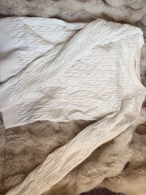 Vit kabelstickad tröja - Säljer en klassisk vit kabelstickad tröja med rund hals och ribbade muddar.  Materialet känns mjukt och bekvämt, och den passar lika bra till jeans som till kjol. Storlek S. 