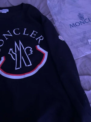 Svart Moncler sweatshirt med logga - Säljer en svart sweatshirt från Moncler med stor vit och röd logga på bröstet. Tröjan har rund halsringning och Moncler-märke på ärmen. Perfekt för dig som gillar streetwear och vill ha något stilrent men ändå iögonfallande.