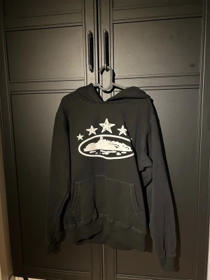 Corteiz HOODIE  - Svart hoodie med stor ficka framtill och huva. På bröstet finns ett vitt tryck med tre stjärnor och ett motiv av ett landskap eller berg. Hoodien har ribbade muddar vid ärmar och nederkant. Perfekt för en avslappnad och cool stil.  HOODIEN är i storlek S men passar även M. Den är i 8/10 skick 