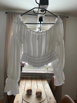 Vit offshoulder blus från Amisu - Supersöt vit offshoulder blus från Amisu med långa puffärmar och volangdetaljer vid midjan och ärmsluten. Blusen har en croppad passform och är perfekt för en trendig look. Tillverkad i ett lätt och luftigt material.