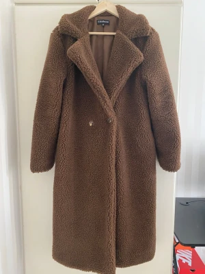 Brun teddyjacka från EllyBazar - Säljer en lång, brun teddyjacka från EllyBazar med bred krage och dubbelknäppning framtill. Jackan har ett mjukt, fluffigt material och är helfodrad på insidan. Perfekt för dig som vill ha en mysig och trendig look under kyliga dagar.
