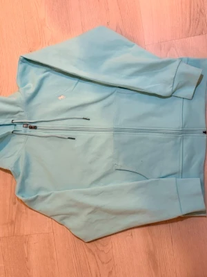 Ljusblå hoodie från Ralph Lauren - Snygg ljusblå hoodie från Ralph Lauren med dragkedja framtill och två fickor. Klassisk broderad logga i rosa på bröstet. Tillverkad i mjukt bomullsmaterial och har justerbar huva med snören. Perfekt för en avslappnad och stilren look.