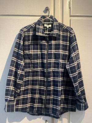 Blårutig overshirt från Kappahl - Overshirt i blått och vitt rutmönster från Kappahl. Modellen har dragkedja framtill, två bröstfickor och klassisk krage. Tillverkad i mjukt tyg som känns som borstad bomull. Perfekt lager på lager-plagg för en avslappnad stil.