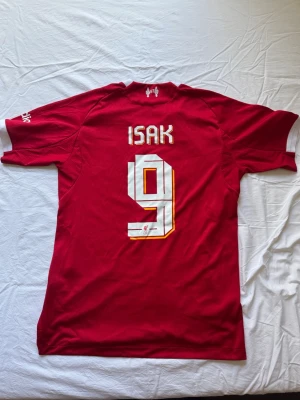 Liverpool matchtröja Alexander Isak - Röd Liverpool FC matchtröja från Adidas med vita detaljer och tryck. Nummer 9 och namnet ISAK på ryggen. Champions League-märke på ärmen och Standard Chartered-logga på bröstet. Spelarverson och material i funktionspolyester. 