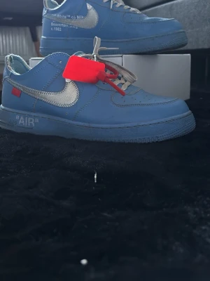 Nike x Off-White Air Force 1 blå - Säljer ett par Nike x Off-White Air Force 1 i blått läder med silverfärgad Swoosh, röda detaljer och den ikoniska röda taggen. Skorna har texten 'AIR' på sulan och tydliga Off-White-signaturer. Perfekta för dig som gillar streetwear och unika sneakers.