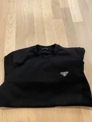 Svart sweatshirt från Prada - En stilren svart sweatshirt från Prada med rund halsringning och lång ärm. Tröjan har en diskret Prada-logga på bröstet och är tillverkad i mjukt material som känns skönt mot huden. Perfekt för en clean och lyxig look.