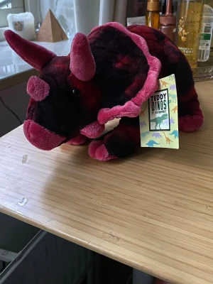 Mjukisdjur Triceratops Teddy Dinos - Säljer ett mjukisdjur i form av en triceratops från Teddy Dinos. Den är gjord i mjuk plysch och har en mix av mörkröda, svarta och rosa färger. Perfekt för dig som gillar dinosaurier och vill ha något mysigt att krama.