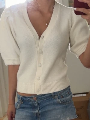 Vit kortärmad kofta med knappar - Snygg vit kofta med korta puffärmar och djup v-ringning. Koftan har ribbade muddar och stängs med stora vita knappar framtill. Perfekt att styla med jeans eller kjol för en fräsch look.