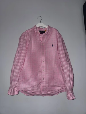 Rosa rutig skjorta från Polo Ralph Lauren - Snygg rosa och vit rutig skjorta från Polo Ralph Lauren med klassisk krage och knappar framtill. Skjortan har lång ärm och ikonisk broderad logga på bröstet. Perfekt för en stilren och avslappnad look.