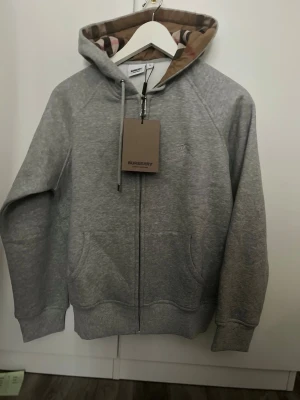 Grå zip-hoodie från Burberry - Snygg grå hoodie med dragkedja från Burberry. Klassisk huva med det ikoniska rutmönstret på insidan, dragsko och två fickor framtill. Tillverkad i mjukt bomullsmaterial, perfekt för en chill och stilren look.