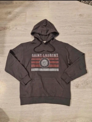 Mörkgrå Saint Laurent hoodie - Snygg mörkgrå hoodie från Saint Laurent med tryck framtill i rött, vitt och svart. Klassisk huva med dragsko och stor magficka. Storlek s-m