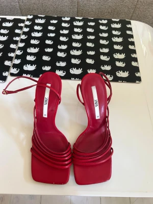 Röda strappy pumps från Zara - Snygga röda pumps från Zara med smala remmar och fyrkantig tå. Skorna har hög klack och en elegant design med ankelrem. Tillverkade i konstläder med glansig finish. Perfekta för att lyfta vilken outfit som helst.