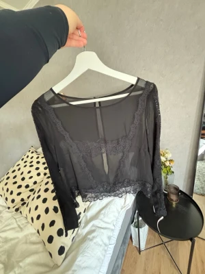 Svart spetstopp  - Snygg svart transparent blus med långa ärmar och fina spetsdetaljer längs halsringning och nederkant. Blusen har en croppad passform och är perfekt för att skapa en trendig lager-på-lager-look.