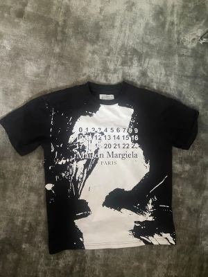 Maison Margiela svartvit t-shirt - Cool svart t-shirt från Maison Margiela med vit abstrakt print och ikonisk logga samt siffror på bröstet. Rund halsringning och korta ärmar. Perfekt för dig som gillar streetstyle och unika designer. 