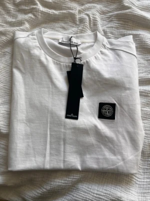 Vit Stone Island t-shirt med logga - Snygg vit t-shirt från Stone Island med klassisk rund hals och korta ärmar. På bröstet sitter den ikoniska svarta logotypen i tygpatch. Tillverkad i mjuk bomull som känns skön mot huden. Perfekt basplagg för en clean och stilren look.