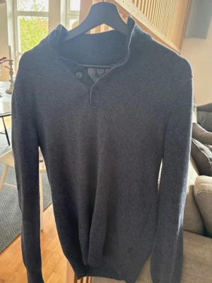 Mörkgrå stickad tröja Stone Island - Mörkgrå stickad tröja från Stone Island med hög krage och två knappar vid halsen. Tröjan har lång ärm och ribbstickade muddar. Perfekt för kyliga dagar och ger en stilren look.