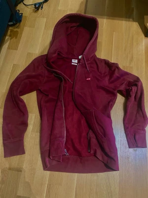 Vinröd hoodie från Levi's - Säljer en vinröd hoodie från Levi's med dragkedja framtill och två fickor. Klassisk huva med snören och liten Levi's-logga på bröstet. Tillverkad i mjukt bomullsmaterial som är skönt att ha på sig. Perfekt för dig som gillar en enkel men snygg stil.