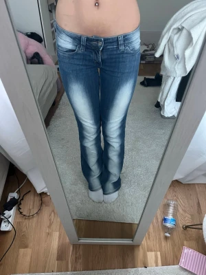 Blå bootcut jeans  - Säljer ett par blå bootcut jeans från cheap monday med snygga slitningar och ljusa partier framtill och baktill. Jeansen har låg midja, klassiska fickor och en stor vit patch med logga bak. Perfekta för en trendig och avslappnad look.