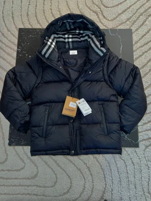 Svart pufferjacka från Burberry - Snygg svart pufferjacka från Burberry med klassiskt rutigt foder i huvan. Jackan har dragkedja, två fickor med dragkedja och en mysig huva. Perfekt för kalla vinterdagar och riktigt trendig look.