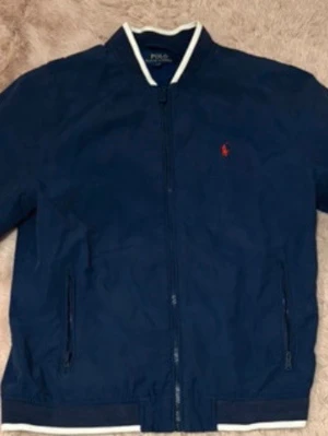 Marinblå bomberjacka Polo Ralph Lauren - Snygg marinblå bomberjacka från Polo Ralph Lauren med vit kontrastkant vid krage och nederkant. Jackan har dragkedja framtill, två fickor med dragkedja och den klassiska röda loggan broderad på bröstet. Perfekt för en stilren och sportig look.