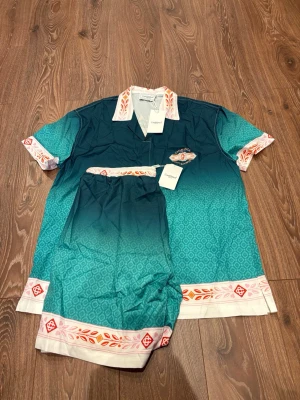 Grön-blå sommardress från Casablanca - Snygg tvådelad sommardress från Casablanca i grön-blått med mönstrade detaljer i vitt, orange och rosa. Skjortan har kort ärm, krage och tryckknappar, samt broderad logga och texten 'Unity is Power' på ryggen. Matchande shorts med elastisk midja. Perfekt för chillkvällar.