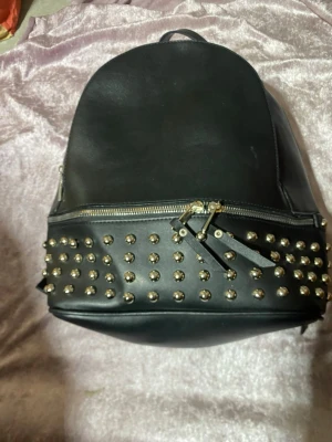 Svart ryggsäck med nitar - Cool svart ryggsäck i konstläder med silvriga nitar på framsidan och dubbla dragkedjor. Justerbara axelband och praktiskt handtag upptill. Perfekt för dig som vill ha en edgy och trendig look.