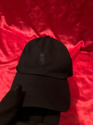 Svart keps från Polo Ralph Lauren - Svart keps från Polo Ralph Lauren med klassisk böjd skärm och broderad logga framtill. Justerbart spänne baktill i metall och broderad POLO-text. Tillverkad i bomull för en bekväm passform.