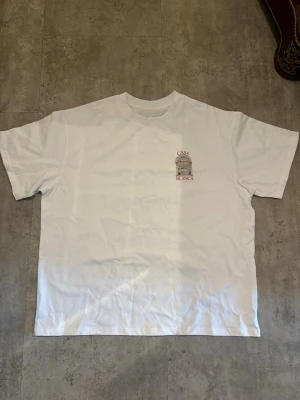 Vit Casa Blanca t-shirt med tryck - Säljer en vit t-shirt från Casa Blanca med ett snyggt tryck av en portal och texten 'Casa Blanca' både fram och bak. T-shirten har korta ärmar och en klassisk passform. Perfekt för dig som gillar stilrena plagg med unik detalj.
