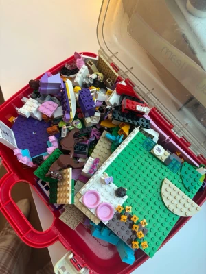 Lego Friends blandade bitar i låda - En röd plastlåda fylld med blandade Lego Friends-bitar i olika färger som lila, rosa, grönt och vitt. Innehåller flera minifigurer, djur, tillbehör och byggplattor. Perfekt för dig som vill bygga egna kreationer eller komplettera din samling.
