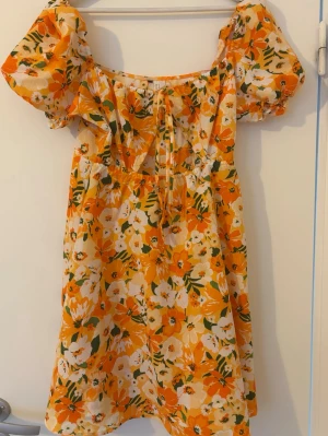 Blommig kortklänning med puffärmar - Superfin kortklänning med blommigt mönster i orange, vitt och grönt. Klänningen har puffiga kortärmar, söt snörning framtill och en luftig passform. Perfekt för varma dagar och ger en somrig vibe. Tillverkad i ett lätt och skönt material.