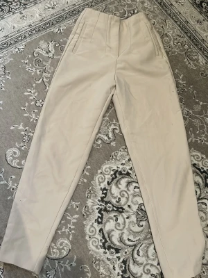 Beige kostymbyxor med hög midja - Snygga beige kostymbyxor med hög midja och raka ben. Byxorna har pressveck framtill och diskreta sidfickor. Perfekta för en clean och stilren look.