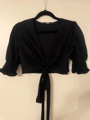 Svart crop top med knytning från Bik Bok - Svart crop top från Bik Bok med djup v-ringning, puffärmar med smock vid ärmslut och snygg knytning framtill genom en ringdetalj. Toppen är i mjukt stretchigt material och har en trendig, figurnära passform.