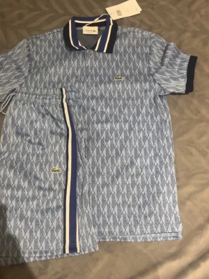 Blått set från Lacoste med mönster - Säljer ett blått set från Lacoste med kortärmad pikétröja och matchande shorts. Setet har ett diskret diamantmönster, mörkblå och vita detaljer på krage och ärmslut samt klassisk Lacoste-logga på både tröja och shorts. Perfekt för en sportig och stilren look.