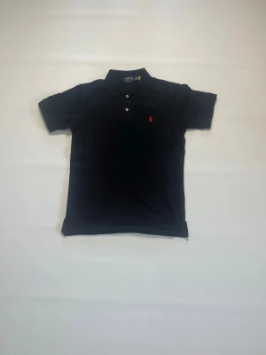 Svart pikétröja från Ralph Lauren storlek M - Klassisk svart pikétröja från Ralph Lauren med korta ärmar och krage. Tröjan har tre knappar framtill och den ikoniska röda loggan broderad på bröstet. Perfekt till jeans eller shorts för en clean look. Pikén är helt ny och aldrig buren och har storlek M