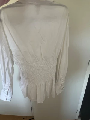 Vit skjorta med smock i midjan - Säljer en vit skjorta från H&M med smockad midja och långa ärmar. Skjortan har klassisk krage, knappar framtill och breda manschetter. Perfekt för dig som gillar en figurnära och trendig look.