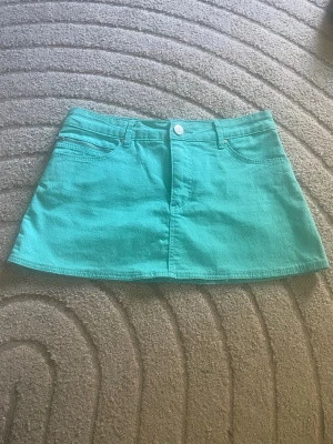 Turkos kortkjol lågmidjad - Snygg lågmidjad turkos kortkjol  med klassisk jeansmodell. Kjolen har fem fickor, bälteshällor och stängs med knapp och dragkedja framtill. Perfekt för dig som vill ha en färgklick i garderoben! midjemått 36 cm(stretchig) längd 29cm