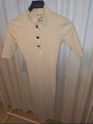 Beige ribbad klänning med knappar - Snygg beige ribbad klänning med kort ärm och hög krage. Klänningen har knappar framtill och är figurnära med en stilren look. Perfekt för dig som gillar enkel men trendig stil. Knappanrna har startat att tappa färg. 