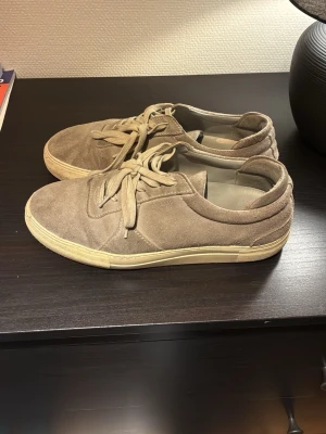 Gråa mockasneakers från Axel Arigato - Snygga sneakers i beige mocka med klassisk låg profil och vita sulor. Skorna har rund tå, ton-i-ton snören och diskret design nött märke. Perfekta för en clean och stilren look till vardagsoutfiten.