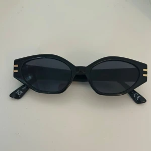 Svarta cat eye solglasögon från Gina Tricot  - Svarta solglasögon i cat eye-modell med mörka glas och gulddetaljer på bågarna från Gina Tricot. 