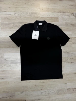 Svart polotopp från Moncler - Svart polotopp från Moncler med klassisk krage och korta ärmar. Diskret Moncler-logga på bröstet och broderad Moncler-text på ena ärmen. Topp i bomull med stilren och modern look, perfekt för dig som gillar exklusiva märken.