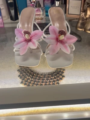Vita pumps med rosa blomdekoration - Unika vita pumps med smala remmar och fyrkantig tå. Framtill sitter stora rosa blommor som ger skorna en riktigt cool och trendig look. Perfekta för dig som vill sticka ut! Klackhöjd är medel och materialet ser ut att vara syntet.