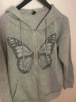 Grå hoodie med fjärilstryck - Grå zip-hoodie med stor glittrig fjäril på framsidan. Hoodien har huva med dragsko, två fickor och dragkedja hela vägen. Perfekt för dig som gillar snygga detaljer och vill sticka ut lite extra.