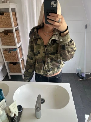 Camouflagemönstrad jacka - Snygg croppad jacka i camouflage-mönster med dragkedja och knappar framtill. Jackan har en loose passform och långa ärmar. Perfekt för dig som vill ha en streetstyle-look.