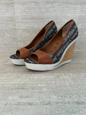 Kilklackar från Calvin Klein - Snygga wedgepumps från Calvin Klein Jeans med öppen tå. Ovandel i brunt läder och svart textil med logotypmönster. Kilklack i kork och vit sula ger en cool kontrast. Perfekta för dig som gillar en trendig och avslappnad stil.
