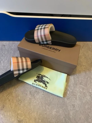 Burberry sandaler med rutigt band - Stilrena sandaler från Burberry med klassiskt beige, svart och rött rutmönster på bandet. Sulan är svart och har en bekväm form. Perfekta för sommaren och ger en lyxig känsla till din outfit. Kommer med originalkartong och dustbag.