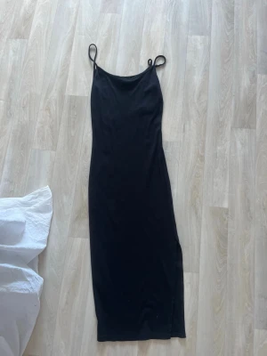 Svart långklänning med smala axelband - En stilren svart långklänning med smala axelband och slits på sidan. Klänningen har en enkel, rak passform och är perfekt för dig som gillar minimalistisk stil. Materialet känns mjukt och följsamt mot kroppen. Slita vid benet 