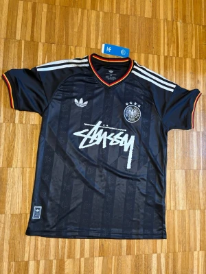 Svart Adidas x Stüssy fotbollströja - Snygg svart fotbollströja från Adidas i samarbete med Stüssy. Tröjan har vita ränder på axlarna, Tysklands landslagsemblem, Stüssy-logga på bröstet och röda detaljer vid krage och ärmslut. Tillverkad i lätt och ventilerande polyester.